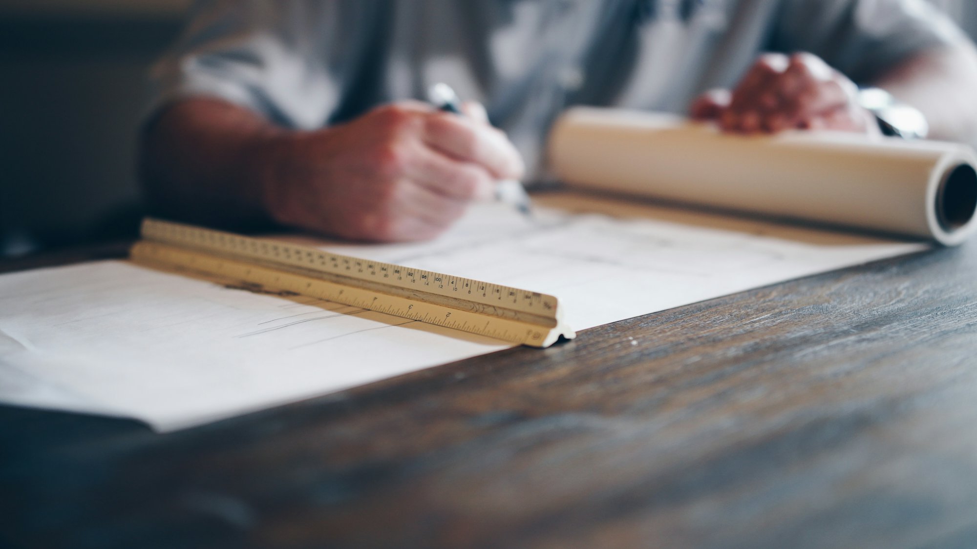 Hands with a pencil over architectural drawings and a scale ruler on a wooden table
